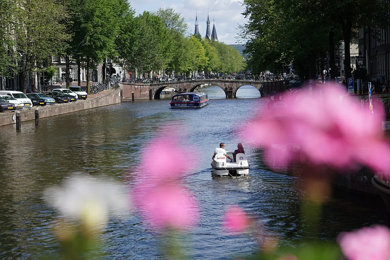 Boats on an Amsterdam canal beneath an arched bridge, with flowers in the foreground